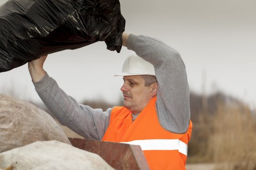 A male waste collection worker wearing a white safety helmet and an orange high-visibility vest is lifting a large black rubbish bag with both hands, preparing to place it into a collection container. The environment appears to be outdoors, possibly on a driveway or a street, with an overcast sky and blurred background of natural landscape. Several other bags of rubbish, primarily made of plastic, are stacked nearby on the ground along with a piece of wooden debris or furniture. The worker's focused expression indicates careful handling of waste, aligning with services offered by Garden Clearance Uxbridge for rubbish removal in the local area. The scene highlights the practical aspects of rubbish collection, emphasizing proper safety gear and handling in a typical outdoor waste management operation.